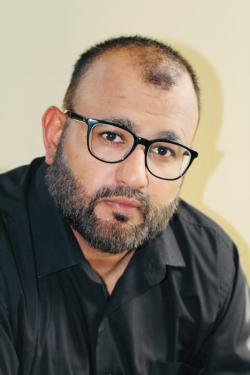 Headshot of Majid Khan in black button-down shirt with black-rimmed glasses against beige background.